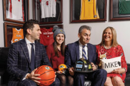 Four people sit together on a couch beneath framed sports jerseys, smiling and holding Milwaukee-themed memorabilia, including a Milwaukee Bucks basketball, a Green Bay Packers helmet, a firefighter’s helmet bearing the Gruber Law Offices logo, and a pillow reading “MKE.”