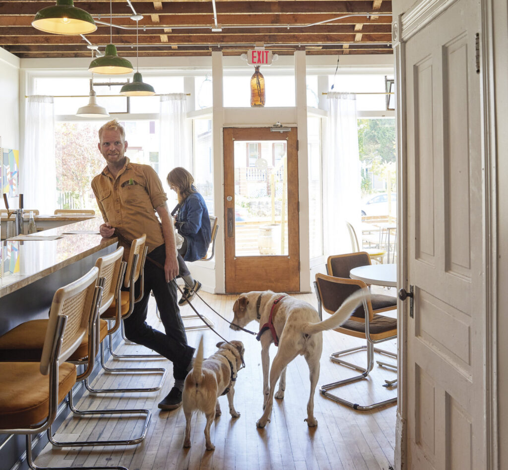 A man in a tan button-down shirt stands beside the bar in a bright, casually furnished neighborhood restaurant as two dogs on leashes greet each other at his feet. A woman sits at the bar behind him, while large front windows, vintage-style pendant lights and wood floors give the space a warm, airy feel.