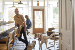 A man in a tan button-down shirt stands beside the bar in a bright, casually furnished neighborhood restaurant as two dogs on leashes greet each other at his feet. A woman sits at the bar behind him, while large front windows, vintage-style pendant lights and wood floors give the space a warm, airy feel.