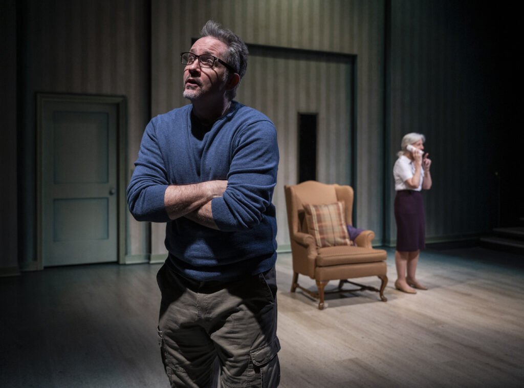 Two people are on a dimly lit stage. A middle aged man in a blue sweater and glasses crosses his arm in the foreground. In the background, an elderly woman stands by an armchair and talks on the phone.