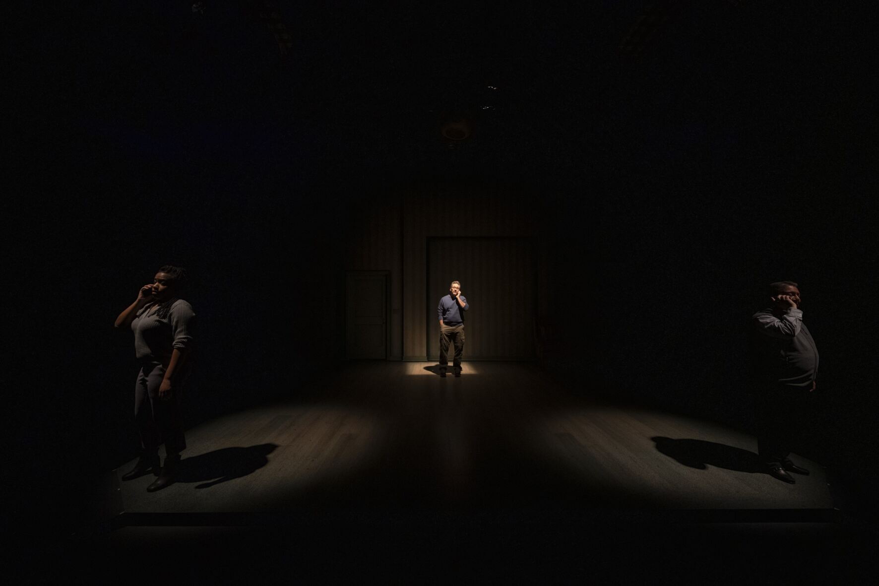 Three people stand on an empty, dark stage with a spotlight on each of them. 
