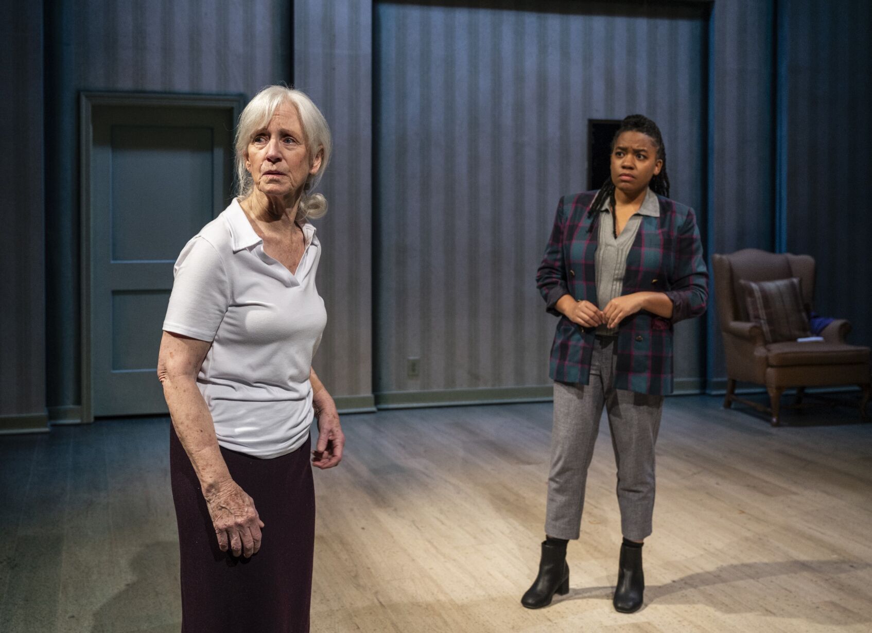 Two women stand on a dimly lit stage. An elderly white woman is in the foreground looking off stage. A young Black woman stands behind her, looking at her. 