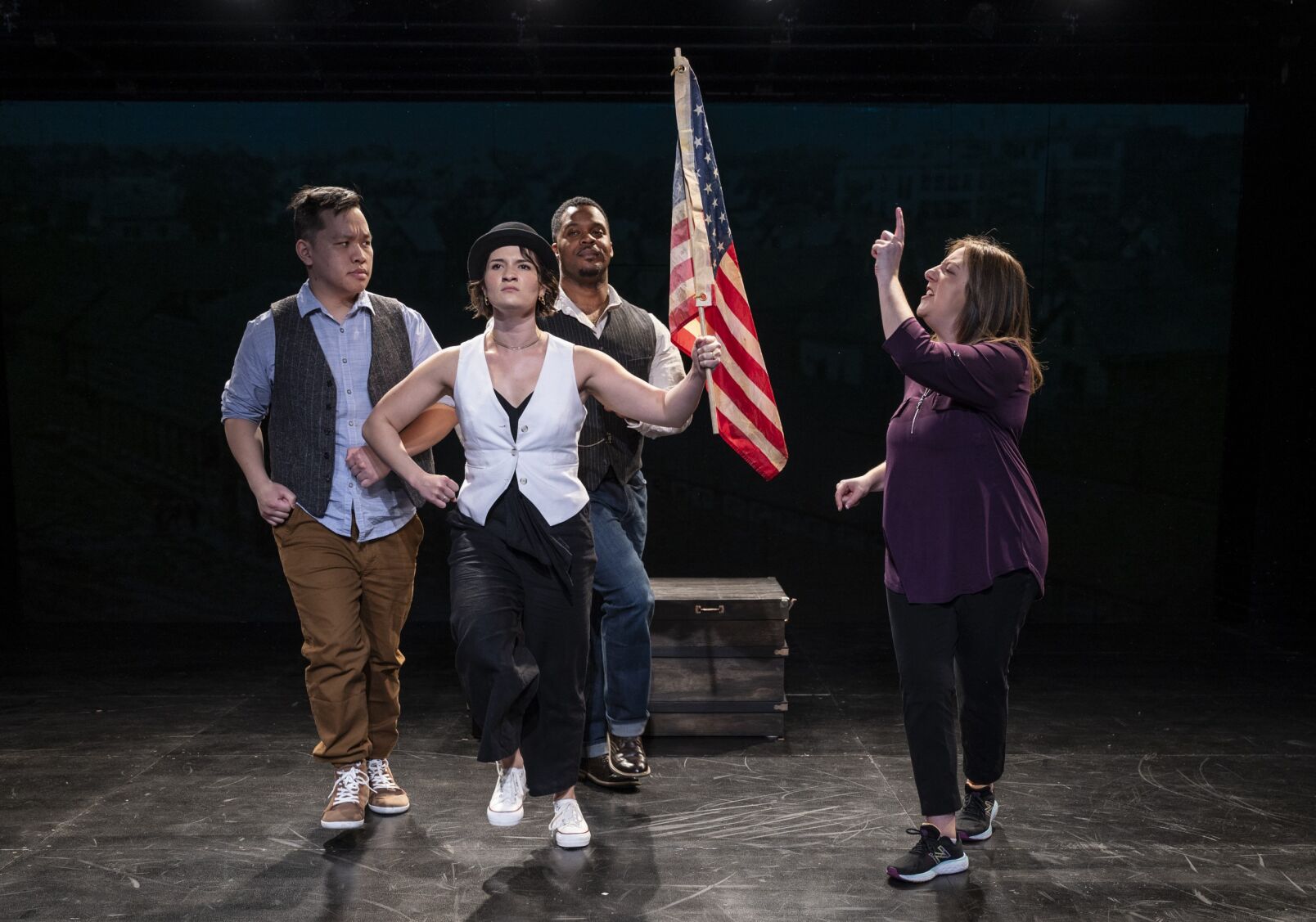 Four actors are on stage in a black box theater. Two men stand in the background. In the foreground, one woman holds an american flag and marches, the other woman yells at her