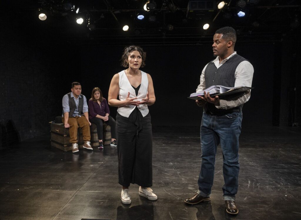 In a black box theater, two actors, a young man and woman stand far in the background. In the foreground, a white woman dressed in pants and a white vest and a black man dressed in jeans and a gray vest, interact with eachother