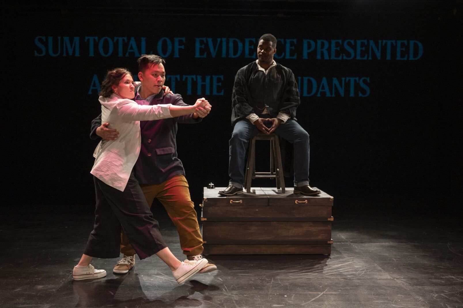Two actors, dance on stage in the foreground, in the background one actor sits on a tall black wearing judges robes.