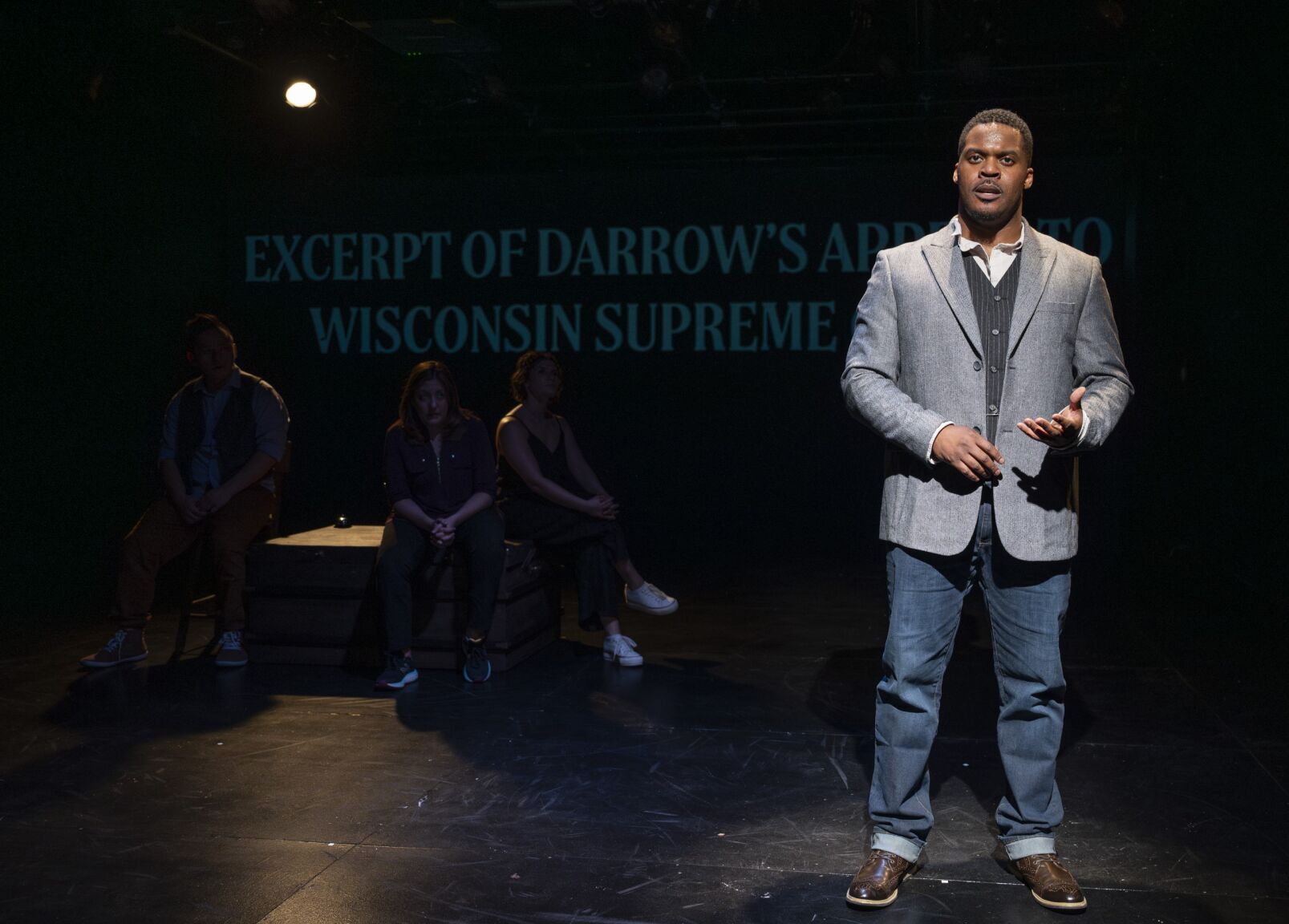 Four actors stand on stage in a black box theater, three far in the background and one young Black actor in jeans and a suit jacket stand off to the right in the foreground.
