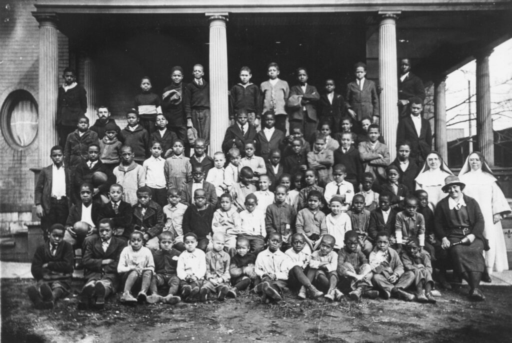 A large group of Black children posing in front of a building with columns, accompanied by priests and nuns at St. Benedict the Moor Parish in Milwaukee, Wisconsin.