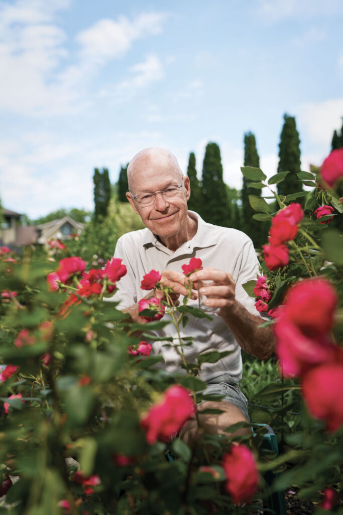 Will Radler, wearing glasses and a white polo, smiles while examining a Knock Out rose in his botanical garden and rosarium.