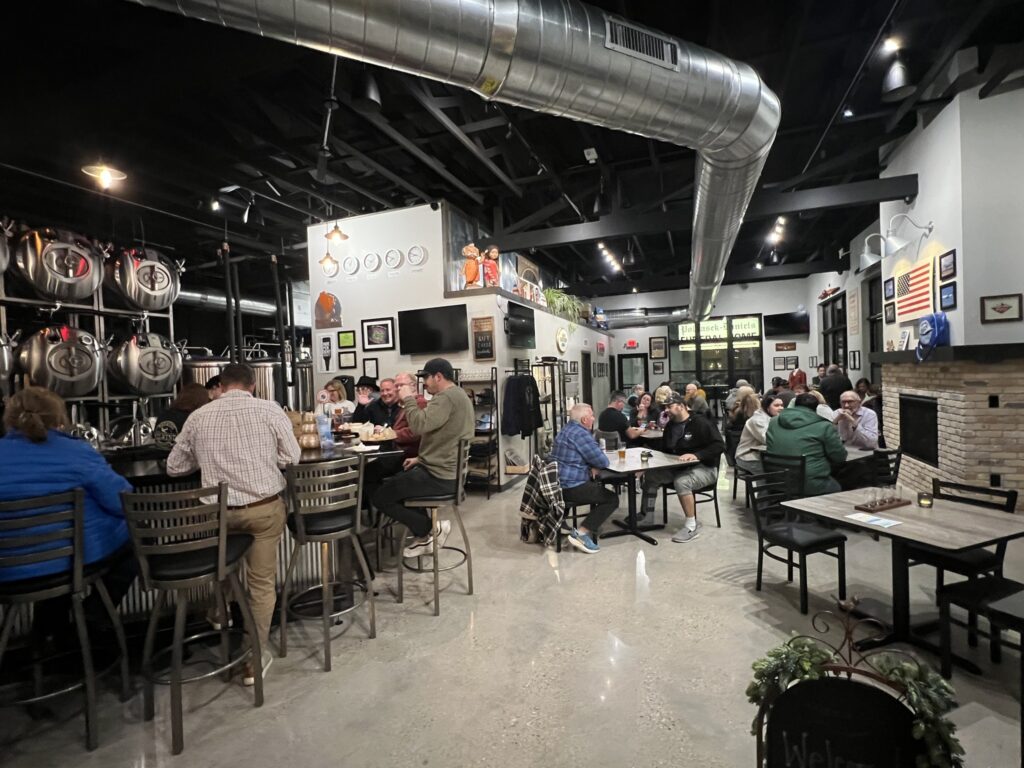 Interior of Tailwinds Brewing filled with people at tables and the bar, enjoying drinks and conversation. Stainless steel brewing tanks line one wall, and décor such as framed art, signs, and an American flag adorn the space. Exposed black ceiling beams and large metal ductwork give the room an industrial feel.