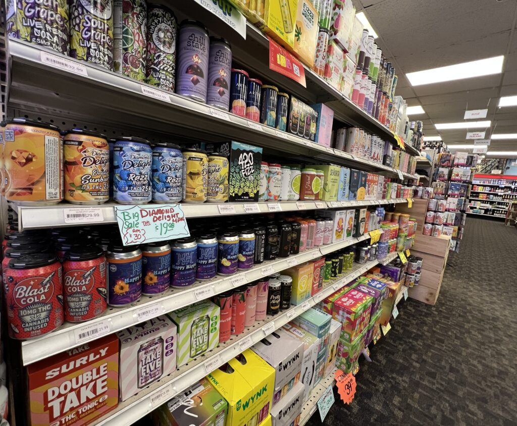 Long retail aisle shot inside Ray's Wine & Spirits in Wauwatosa, WI, showing four large shelves fully stocked with hundreds of brightly colored, branded cans of non-alcoholic and THC-infused beverages, including 'Blast Cola' and 'Big Diamond Delight.'