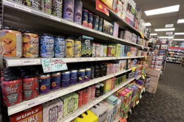 Long retail aisle shot inside Ray's Wine & Spirits in Wauwatosa, WI, showing four large shelves fully stocked with hundreds of brightly colored, branded cans of non-alcoholic and THC-infused beverages, including 'Blast Cola' and 'Big Diamond Delight.'