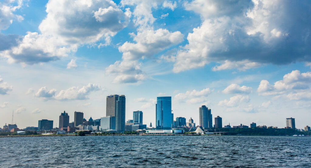 Milwaukee's downtown skyline rises above Lake Michigan beneath a dramatic sky filled with large white cumulus clouds, with the Milwaukee Art Museum and other waterfront landmarks visible along the shoreline.