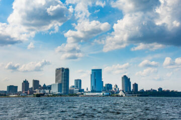 Milwaukee's downtown skyline rises above Lake Michigan beneath a dramatic sky filled with large white cumulus clouds, with the Milwaukee Art Museum and other waterfront landmarks visible along the shoreline.