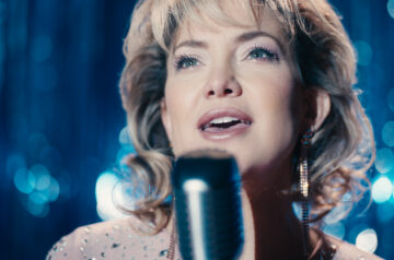 A glamorous female singer performs onstage in close-up, singing into a microphone under cool blue stage lighting, with sparkling bokeh lights behind her; she wears a shimmering dress, dangling earrings, and styled blonde hair, her expression focused and emotional.