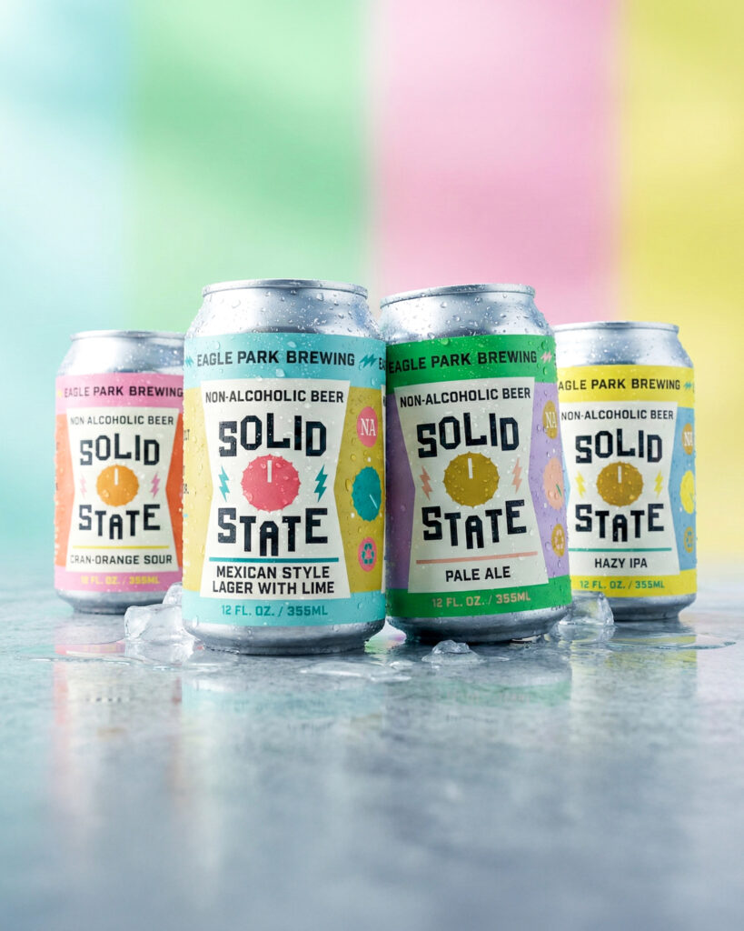 Four chilled cans of Eagle Park Brewing’s Solid State non-alcoholic beer series—Mexican-style lager with lime, pale ale, hazy IPA, and cranberry-orange sour—arranged on ice with visible condensation, set against a softly blurred pastel backdrop divided into vertical bands of mint green, pale pink, light yellow, and sky blue.