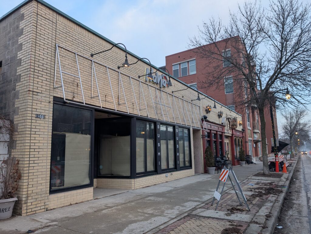 A vacant storefront with paper-covered windows on Oakland Avenue in Shorewood, Wisconsin, marked as the future site of a new pizzeria. The tan brick building sits along a quiet street with neighboring restaurants, bare trees and light construction signage visible on the sidewalk.