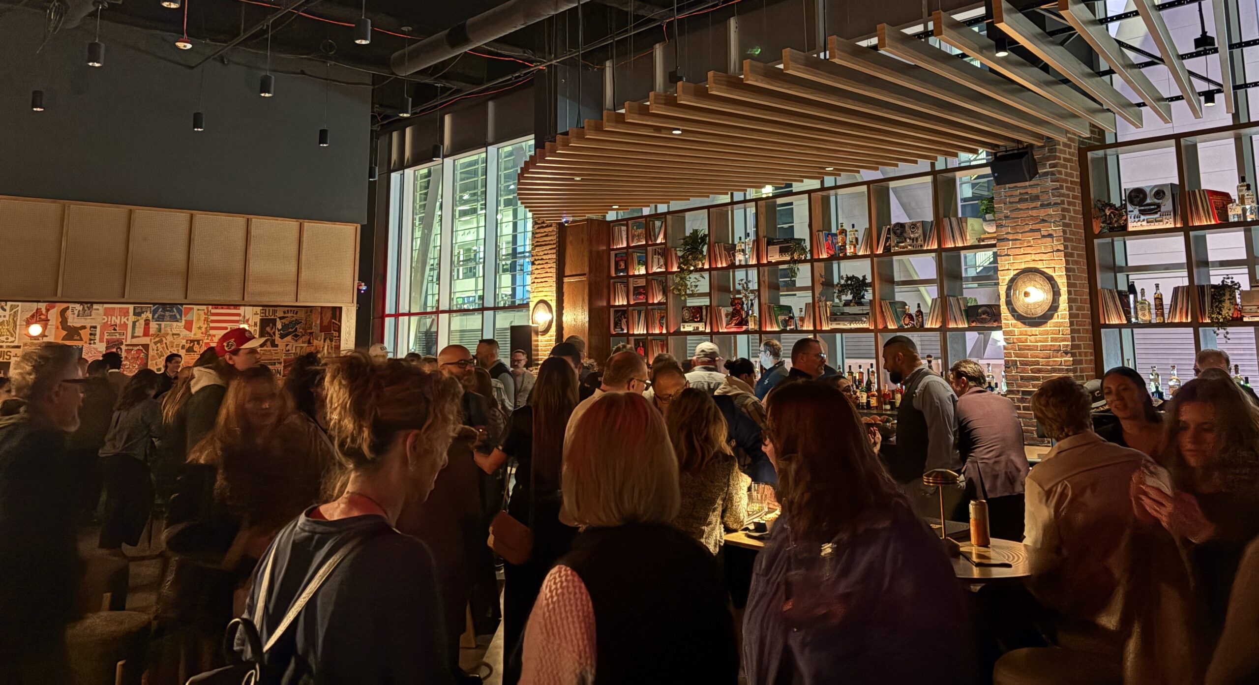 A crowded, warmly lit bar inside a modern industrial space, where guests mingle around high-top tables and a long bar backed by brick walls and shelves filled with records and bottles. Wooden slats hang from the ceiling, and large windows line the room, creating an energetic, social atmosphere.