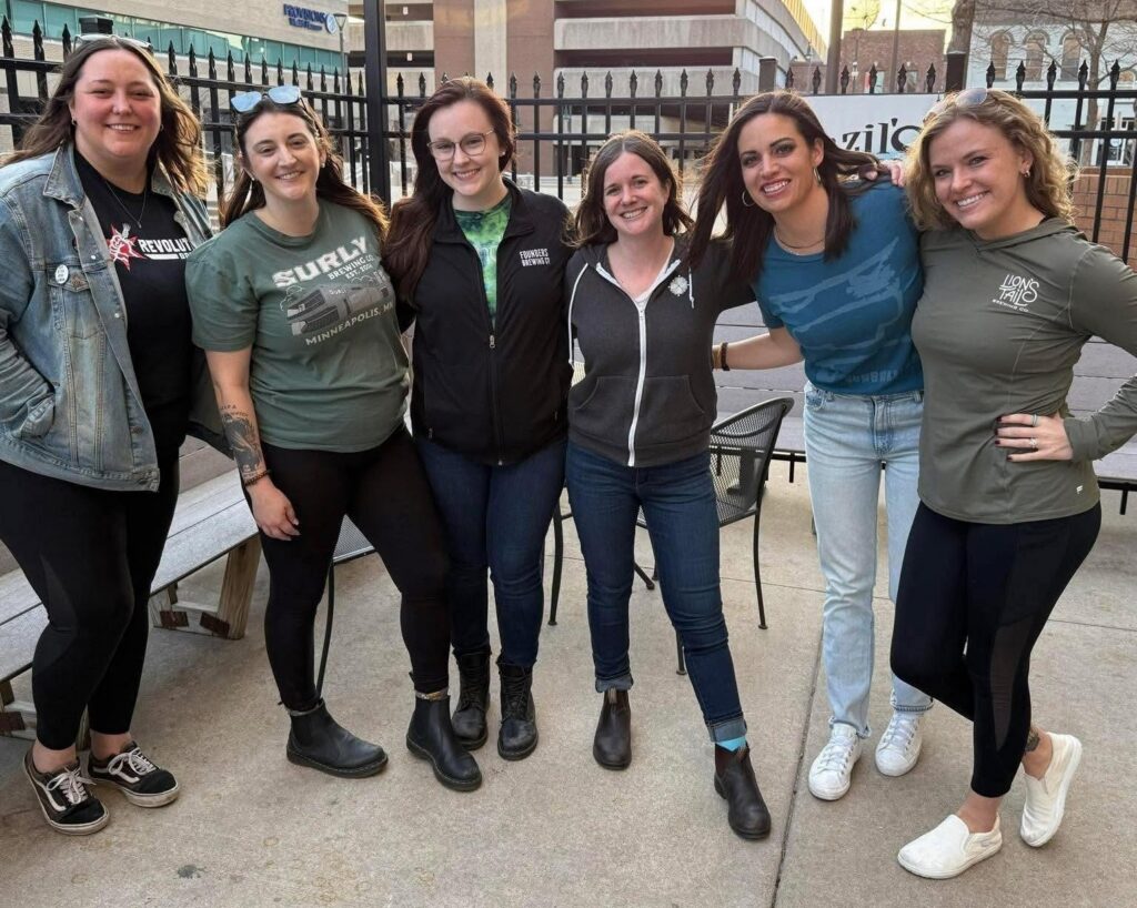Six members of the Women's Beer and Spirits Collective stand arm in arm outdoors on a patio, smiling at the camera. They wear casual brewery-branded shirts and jackets, with city buildings and a fenced sidewalk visible behind them.