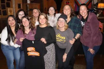 Nine members of the Women’s Beer and Spirits Collective pose together inside a cozy bar, smiling and standing arm in arm, with two holding up a black T-shirt that reads “Her Tomorrow.”