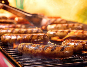 Several sausages sizzle on a hot grill, marked with dark char lines as flames flare up beneath them and metal tongs hover above, turning one over amid rising smoke.