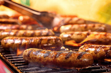 Several sausages sizzle on a hot grill, marked with dark char lines as flames flare up beneath them and metal tongs hover above, turning one over amid rising smoke.