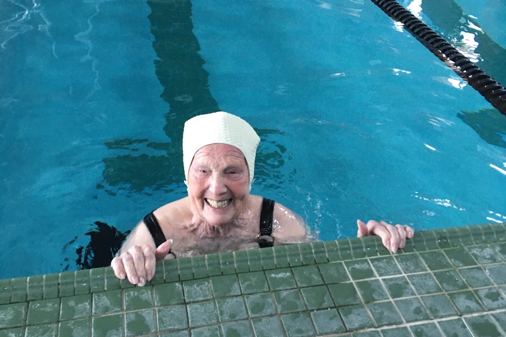 100-year-old swimmer Jean Toepfer smiles in the water at the edge of the pool.