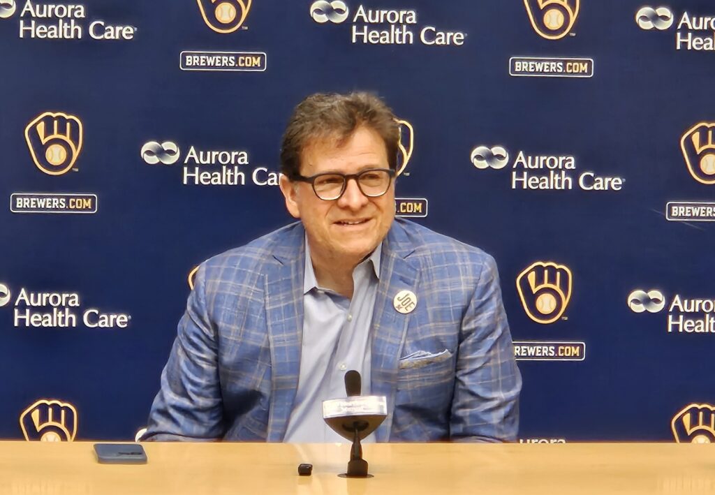 Man in glasses and a blue plaid blazer speaks at a Milwaukee Brewers press conference, seated behind a microphone with a Brewers and Aurora Health Care backdrop.