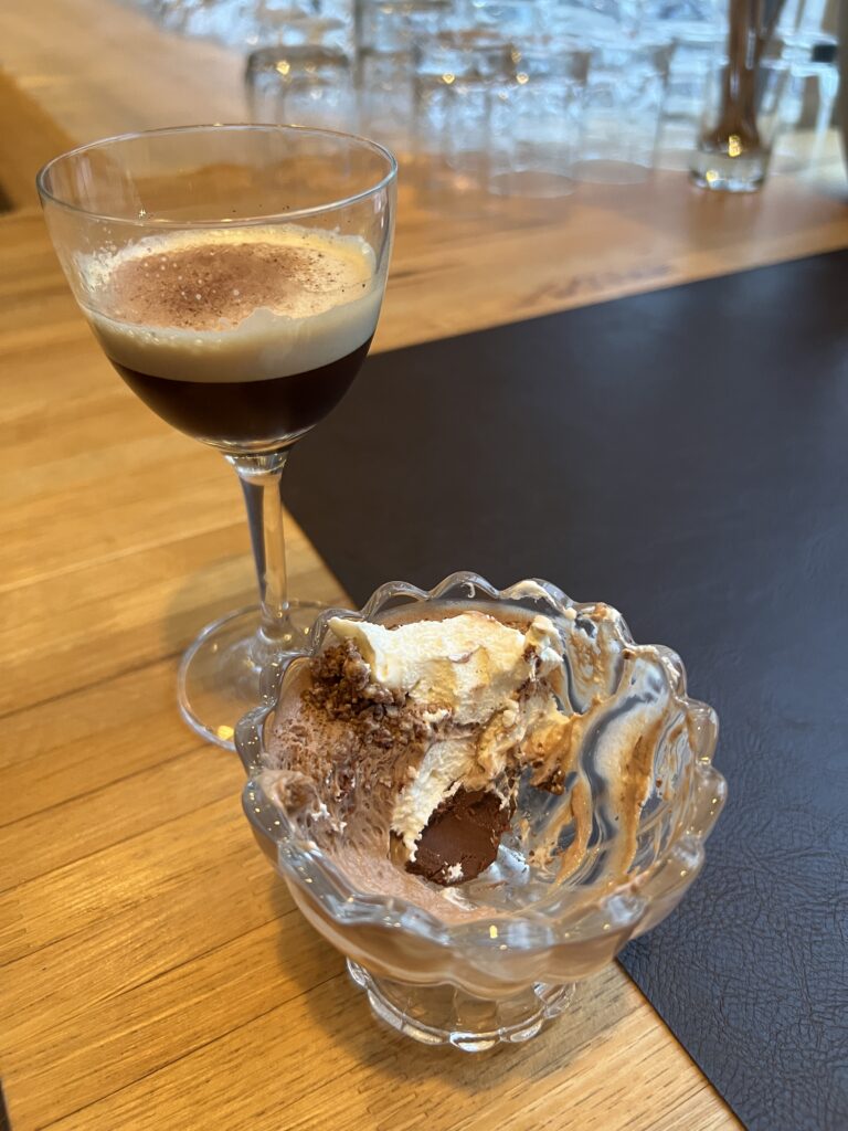 A small glass dish with a partially eaten chocolate parfait topped with white chocolate mousse and crunchy crumbles sits on a wooden bar next to a stemmed glass of dark espresso drink with tan foam and a brown powder on top.