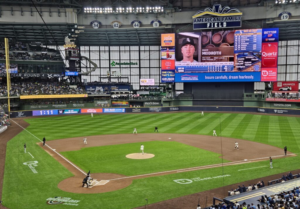 Wide view of American Family Field during a Milwaukee Brewers game, with players in position on the field and a large scoreboard displaying player information.