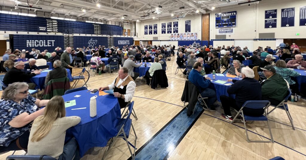 Players fill a high school gym during the Glendale Open Sheepshead tournament, sitting at blue-covered round tables and playing cards beneath “Nicolet Knights” bleachers and a scoreboard.