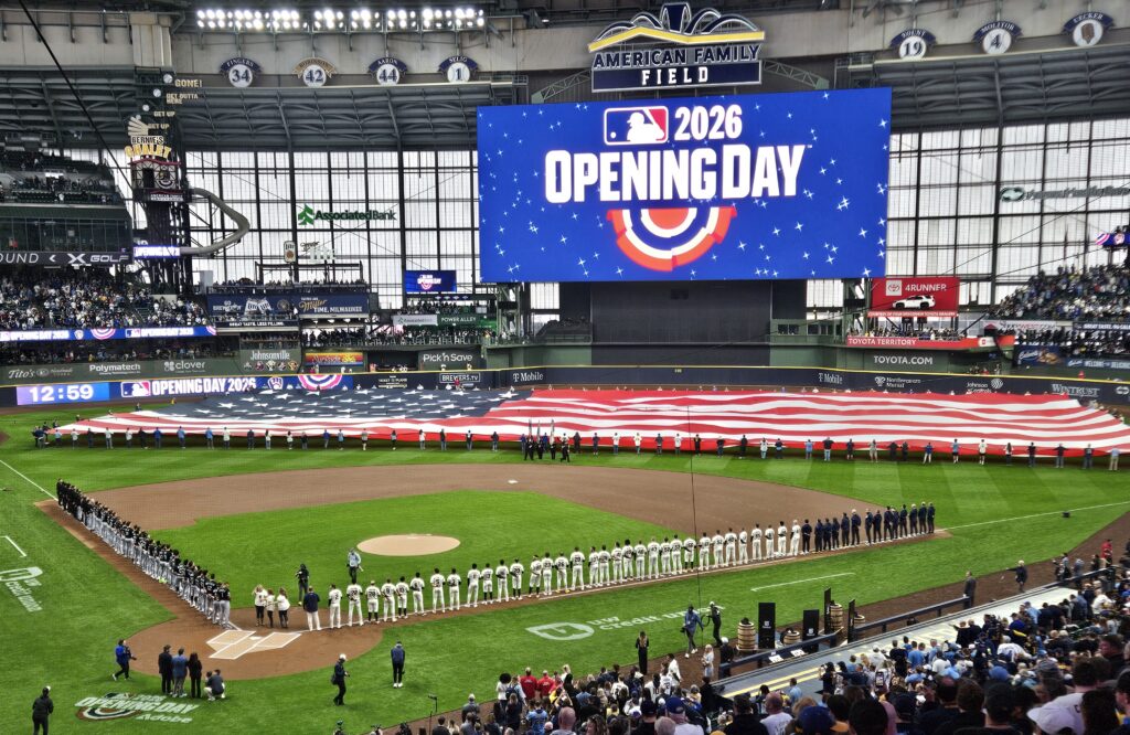 Players line the field during pregame ceremonies at American Family Field as a giant American flag is held across the outfield beneath a scoreboard reading “2026 Opening Day.”