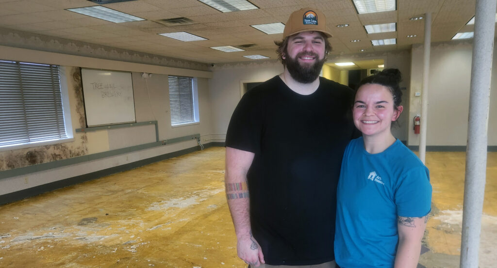 A bearded man and a woman stand smiling inside a partially gutted commercial space, with bare floors and walls behind them and a whiteboard reading “Tree Line Brewery.”
