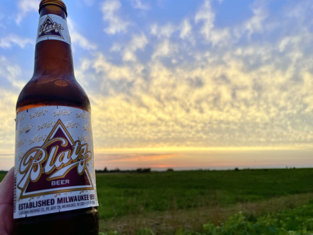A bottle of Blatz beer with a white and gold label reading “Blatz Beer” and “Established Milwaukee 1851” is held up against a wide rural landscape at sunset, with a colorful sky and open fields stretching to the horizon.