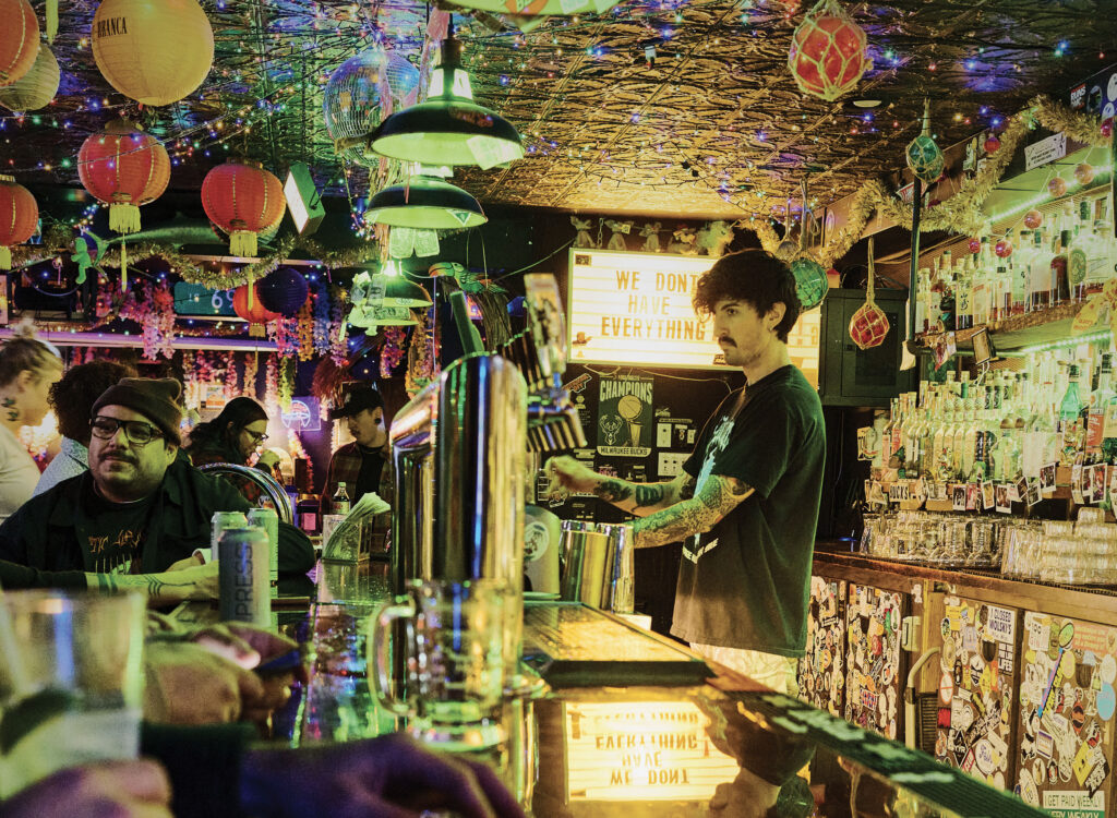 A tattooed bartender pours a drink at a crowded, neon-lit bar decorated with colorful lanterns, string lights, and hanging ornaments, with shelves of liquor bottles behind him and patrons seated along a sticker-covered counter; a lighted sign in the background reads “We don’t have everything.”