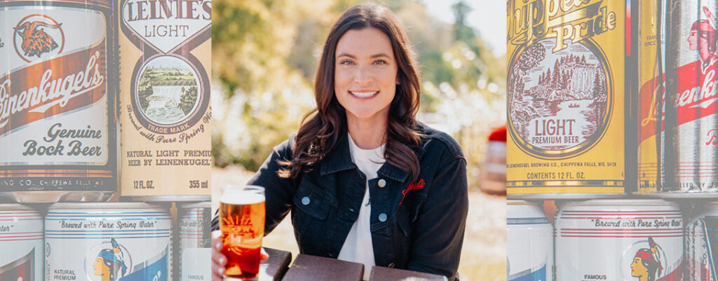 A woman in a black jacket sits outdoors holding a glass of beer, framed by large vintage-style Leinenkugel’s beer can graphics featuring labels for Leinenkugel’s Light and Chippewa Pride Light Premium Beer.