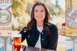 A woman in a black jacket sits outdoors holding a glass of beer, framed by large vintage-style Leinenkugel’s beer can graphics featuring labels for Leinenkugel’s Light and Chippewa Pride Light Premium Beer.