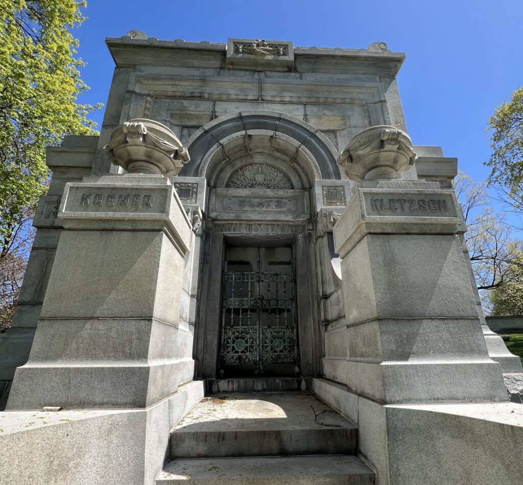 A large stone mausoleum with an arched entrance and ornate metal doors stands in a cemetery, with carved names "Blatz" above the middle door and “Kremer” and “Kletzsch” on the front pillars and trees framing the structure under a clear blue sky.
