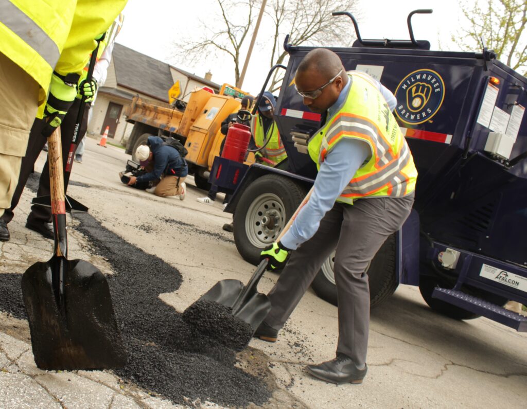 Milwaukee Mayor Cavalier Johnson, wearing business clothes and dress shoes with a reflective safety vest, shovels fresh asphalt into a pothole on a residential street beside a truck with a Milwaukee Brewers logo, as a photographer crouches nearby.