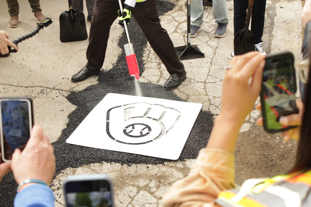 Brewers executive Rick Schlesinger, wearing black dress loafers and business attire, uses a spray can and stencil to paint a Milwaukee Brewers glove logo onto a freshly filled pothole while onlookers record the moment on their phones.