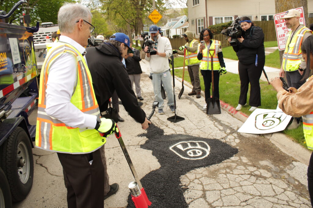 City workers and media gather around a patched pothole on a neighborhood street as a Brewers logo is stenciled onto the new asphalt, with cameras and shovels visible.