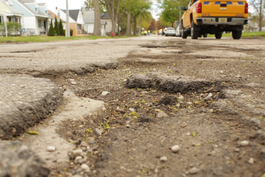 Close-up of a rough, damaged residential street surface with multiple potholes and crumbling asphalt, with houses and parked vehicles blurred in the background.