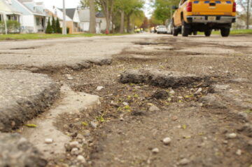 Close-up of a rough, damaged residential street surface with multiple potholes and crumbling asphalt, with houses and parked vehicles blurred in the background.