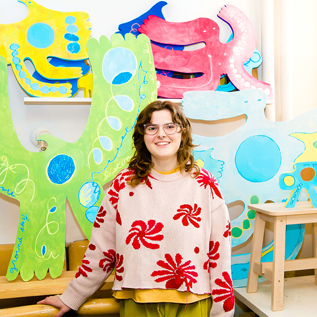 Artist Hattie Grimm poses with colorful painted wooden art of birds with human forms.