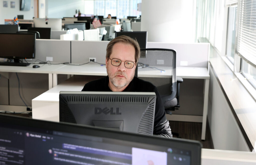 Man wearing glasses works at a desktop computer in a bright, open-plan office with rows of empty cubicles and monitors behind him.