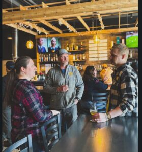 Three people stand and chat at a bar inside a warmly lit brewery taproom, with beer taps, bottles, and TV screens visible behind them.