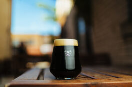 A glass of dark stout beer with a thick foamy head sits on a wooden table, with a softly blurred outdoor setting in the background.