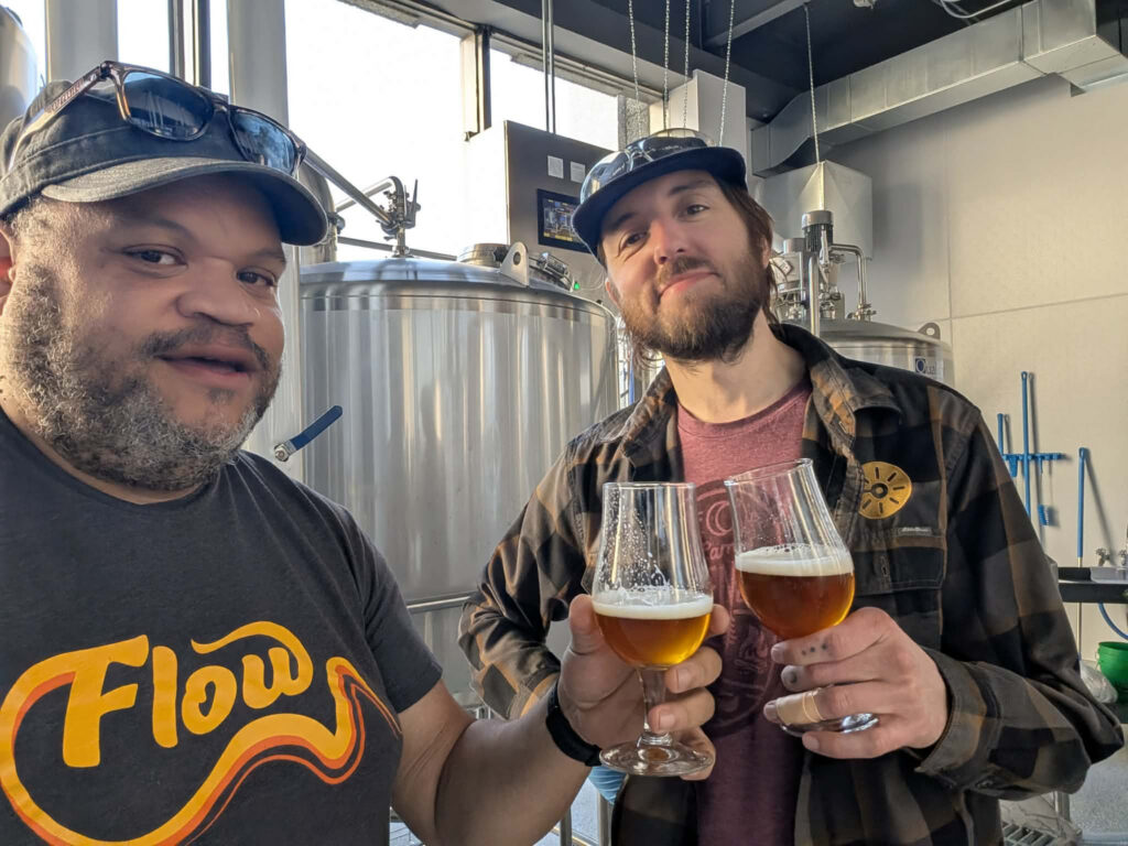 Two men stand inside a brewhouse holding tulip glasses of amber beer for a toast, with stainless steel brewing tanks and equipment in the background.