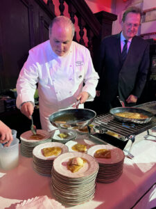Chef in a white jacket prepares plated desserts at a station, spooning sauce over crepes while another man in a suit looks on.