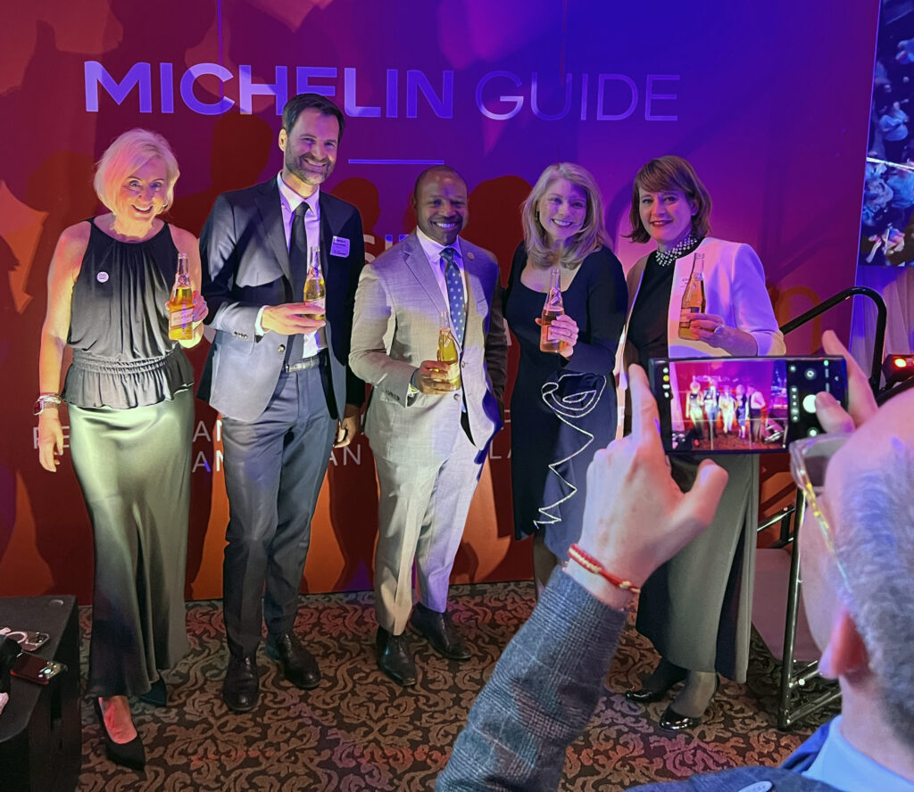 Group of five people pose with clear bottles of Miller High Life in front of a backdrop reading “Michelin Guide,” as someone photographs them with a smartphone in the foreground.