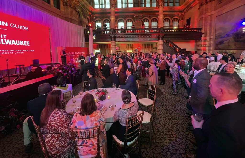 Large crowd gathers in an ornate hall for a Michelin Guide event, with guests seated and standing around tables facing a stage lit in red.